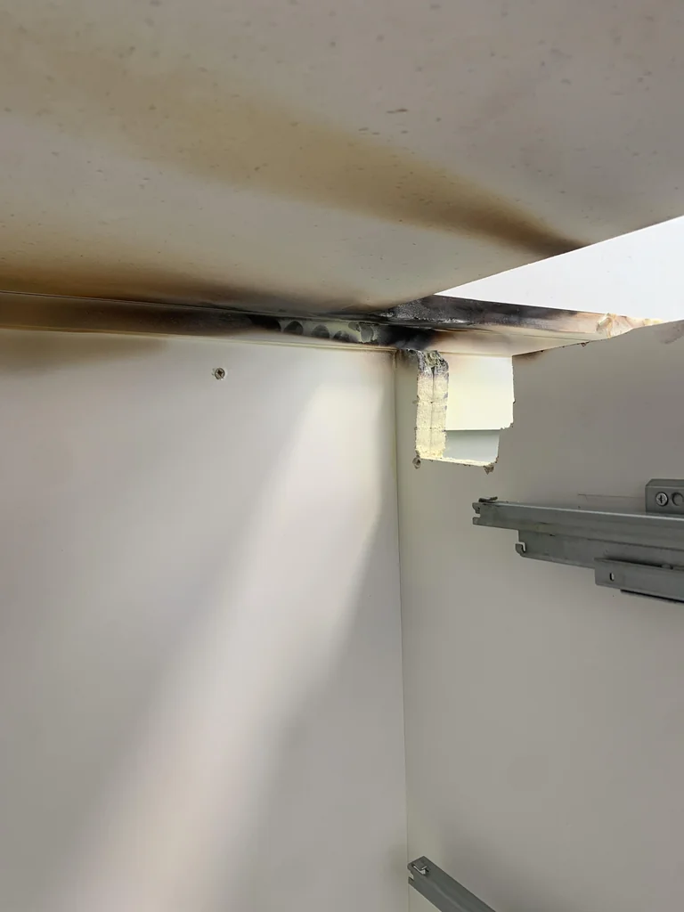 Dark residue and damaged material visible beneath a kitchen benchtop inside open cabinetry