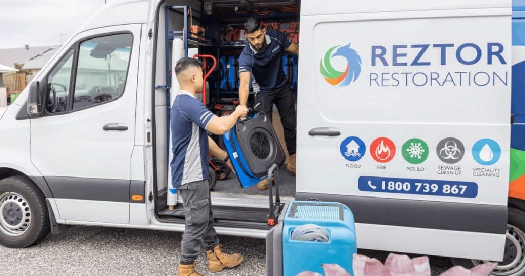 Two Reztor technicians unloading an air mover and dehumidifier from a branded service van on a driveway